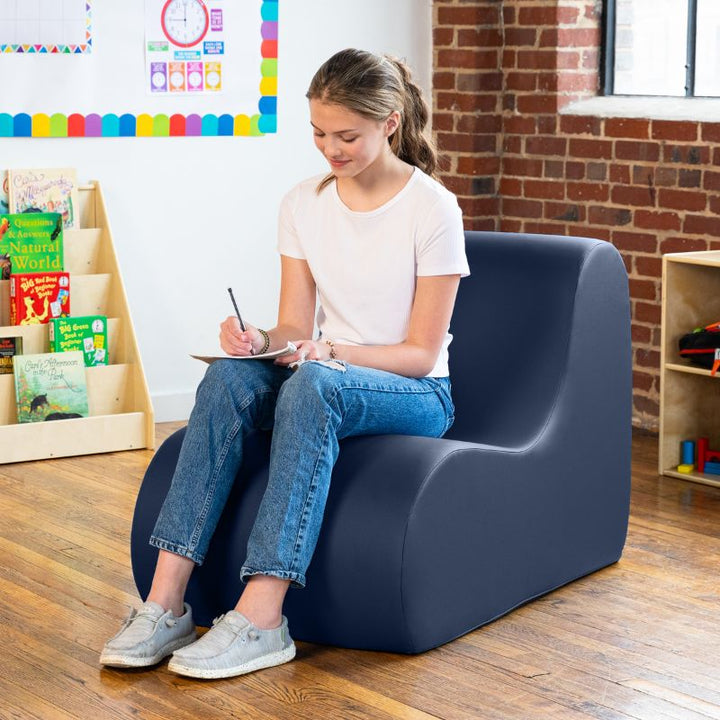 Student sitting in a navy blue Jaxx Midtown foam chair while writing, a wipeable classroom soft seating chair for kids and adults