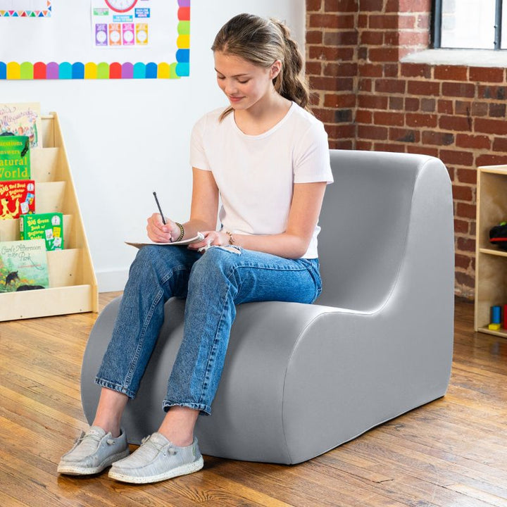 Student sitting in a submarine grey Jaxx Midtown foam chair while writing, a wipeable classroom soft seating chair for kids and adults