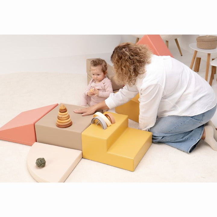 Compact layout of the IGLU Foam Play Set - Creativity with a seated baby and a caregiver arranging toys on the foam climbing blocks