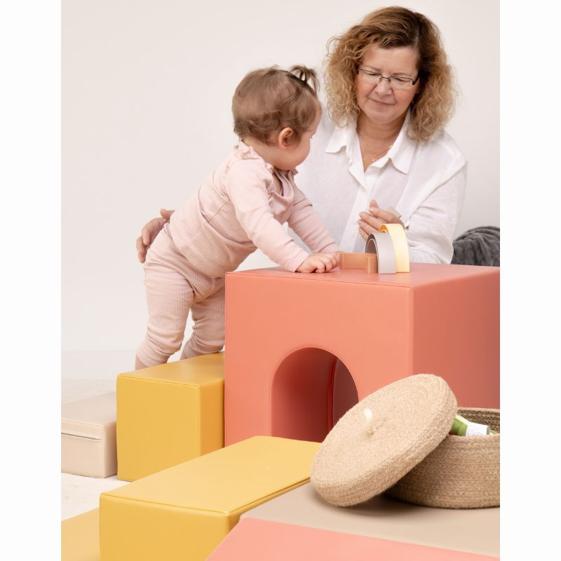 Close-up of a toddler standing at the IGLU Foam Play Set - Creativity tunnel cube, practicing fine motor skills with a rainbow stacker