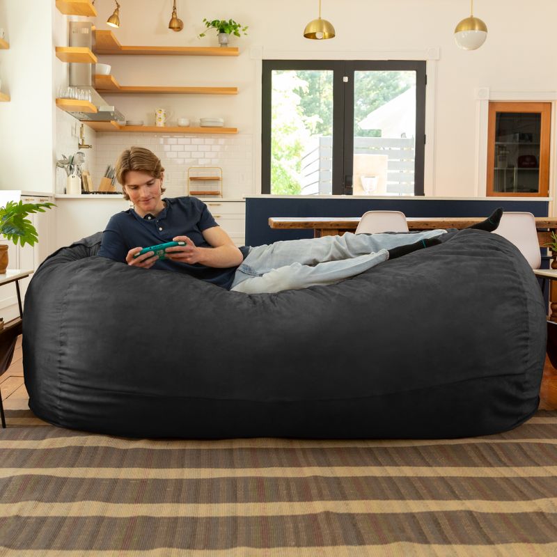 Man playing a handheld game on the Jaxx 7 ft giant bean bag chair in black microsuede in a styled living space
