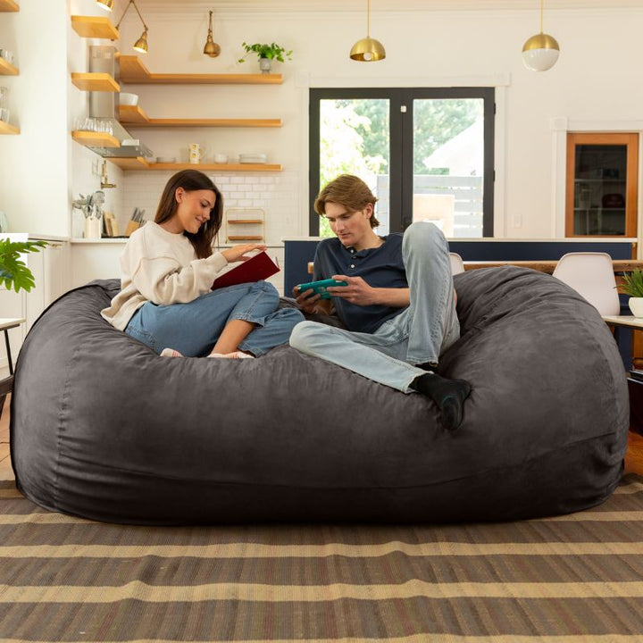 Couple reading and gaming together on the Jaxx 7 ft giant bean bag chair in charcoal microsuede in a styled living space