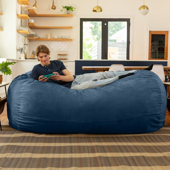 Man playing a handheld game on the Jaxx 7 ft giant bean bag chair in navy microsuede in a styled living space