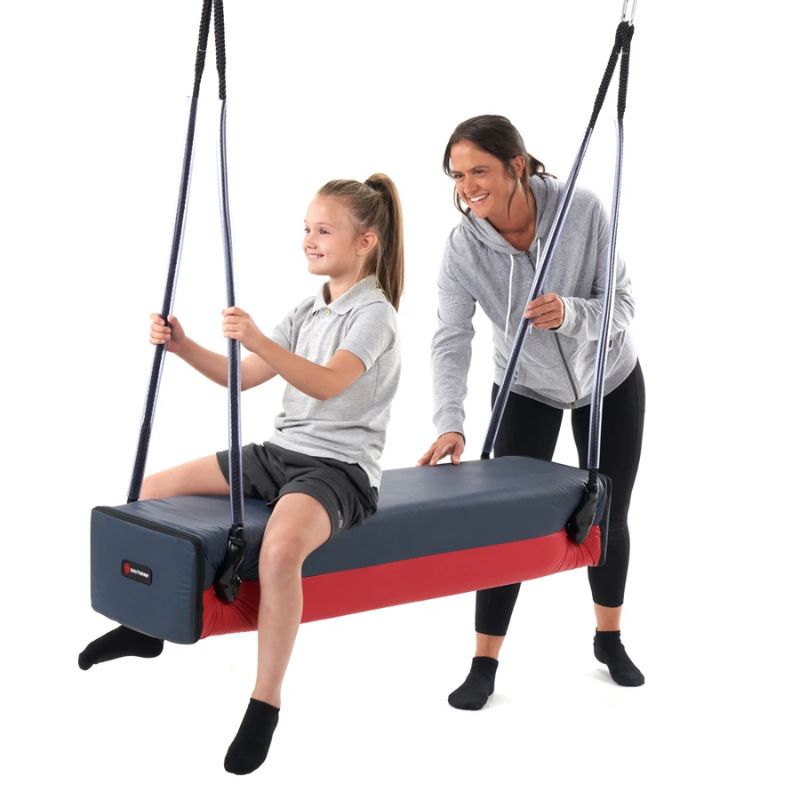 A smiling child holds the ropes of the Southpaw Glider Bolster Swing as a therapist steadies the padded therapy swing beside her