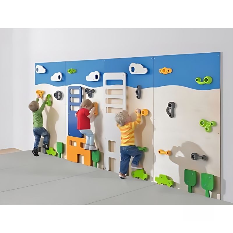 Three children traversing the HABA Pro Climbing World City Wall Panels in a school gym, showing the full four-panel bouldering wall set with colourful birch wood city-themed applications
