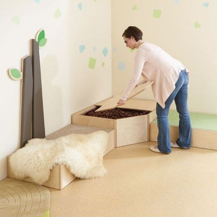 Adult opening the two-piece birch wood lid of the HABA Pro Grow.upp sensory bath platform to reveal chestnut fill, shown in a calm corner classroom setting