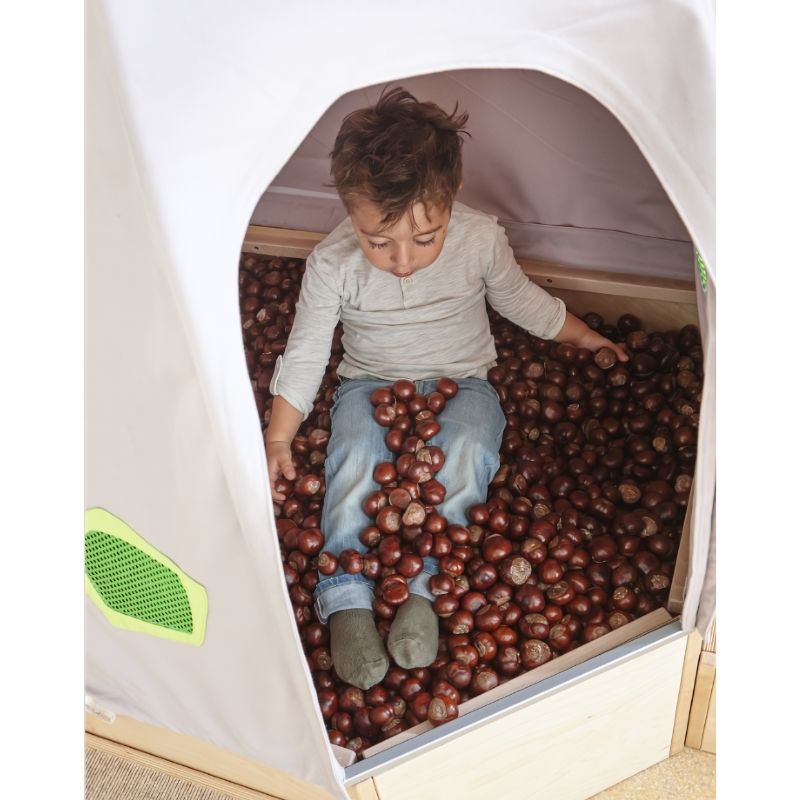 Child sitting in the open HABA Pro Grow.upp sensory bath platform filled with chestnuts for tactile sensory play, viewed through the corner cave entrance
