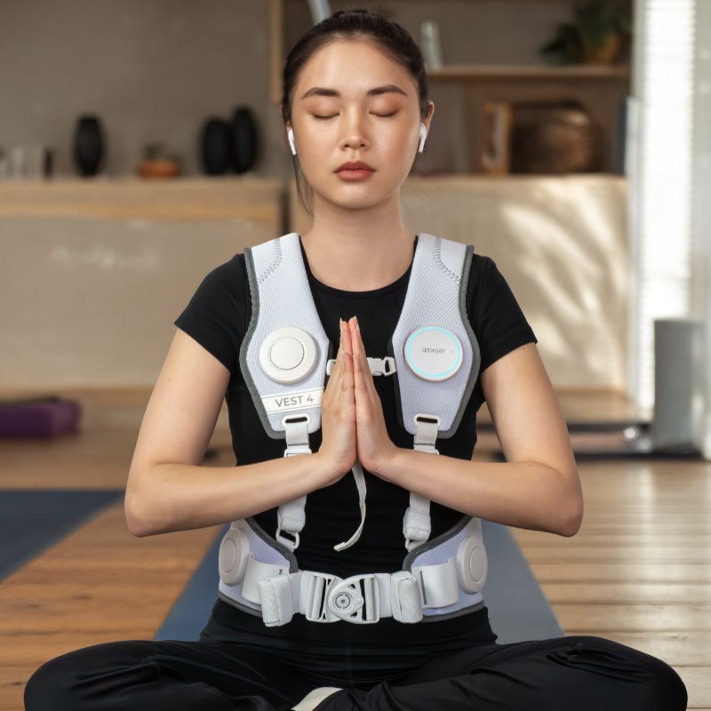 Woman meditating on a yoga mat with eyes closed while wearing the white Woojer Vest 4 haptic vest for vibroacoustic relaxation and breathwork