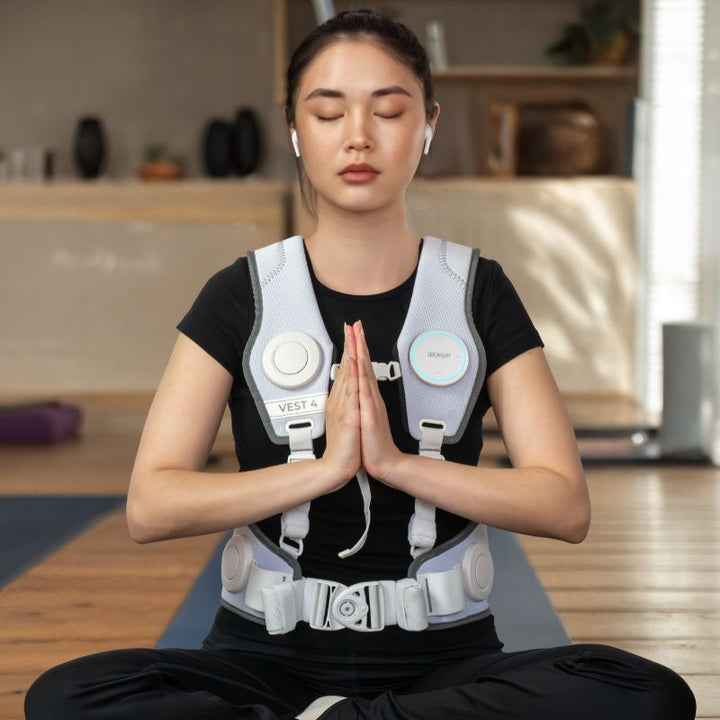 Woman meditating on a yoga mat with eyes closed while wearing the white Woojer Vest 4 haptic vest for vibroacoustic relaxation and breathwork