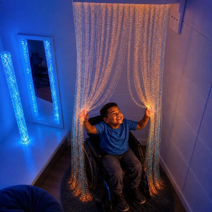 Smiling boy using a wheelchair parting the glowing amber strands of the TFH Wall Mounted Fiber Optic Curtain, a touchable fibre optic sensory light for all abilities