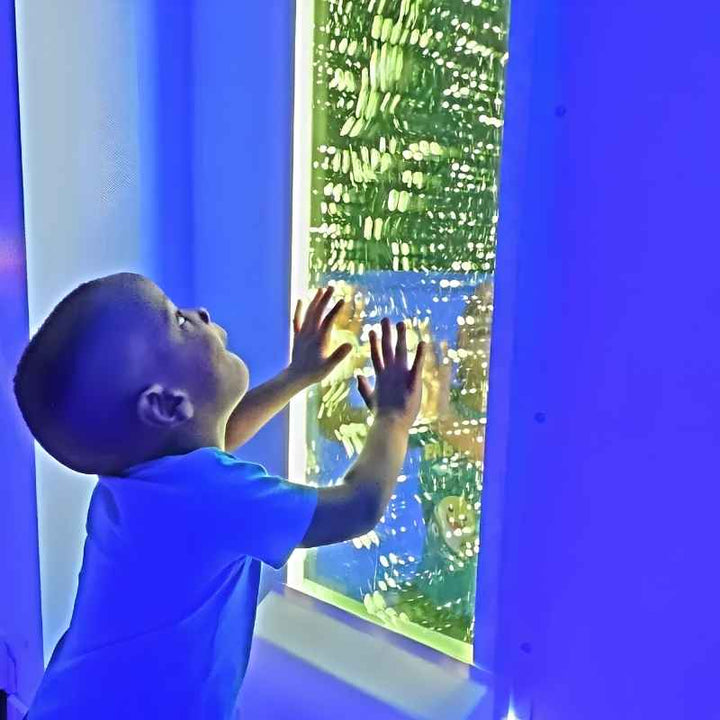 A young child reaches up to touch the glowing green TFH Infinity Bubble Wall, a wall-mounted sensory bubble wall for visual stimulation