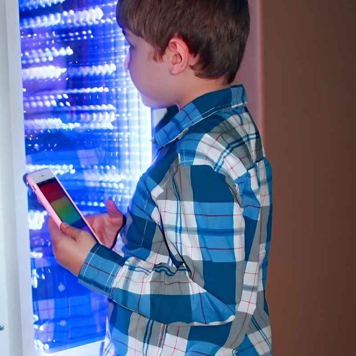 A boy uses the MSE Genie App on his phone to change colours on the blue lit TFH Infinity Bubble Wall, an interactive sensory lighting wall