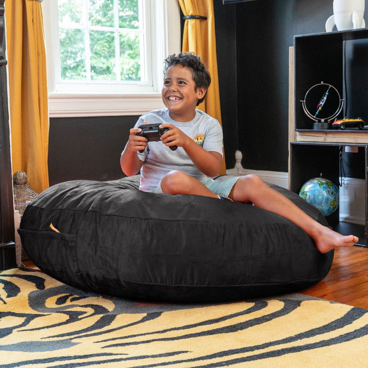 A boy playing video games on a black Jaxx Cocoon 4ft kids bean bag chair, a versatile sensory seat