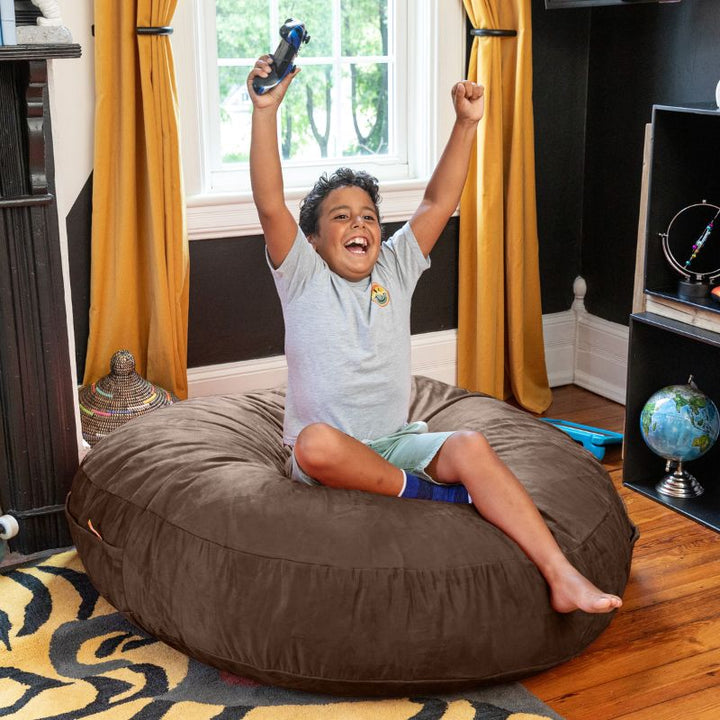 A boy playing video games on a chocolate Jaxx Cocoon 4ft kids bean bag chair, a cozy sensory crash pad