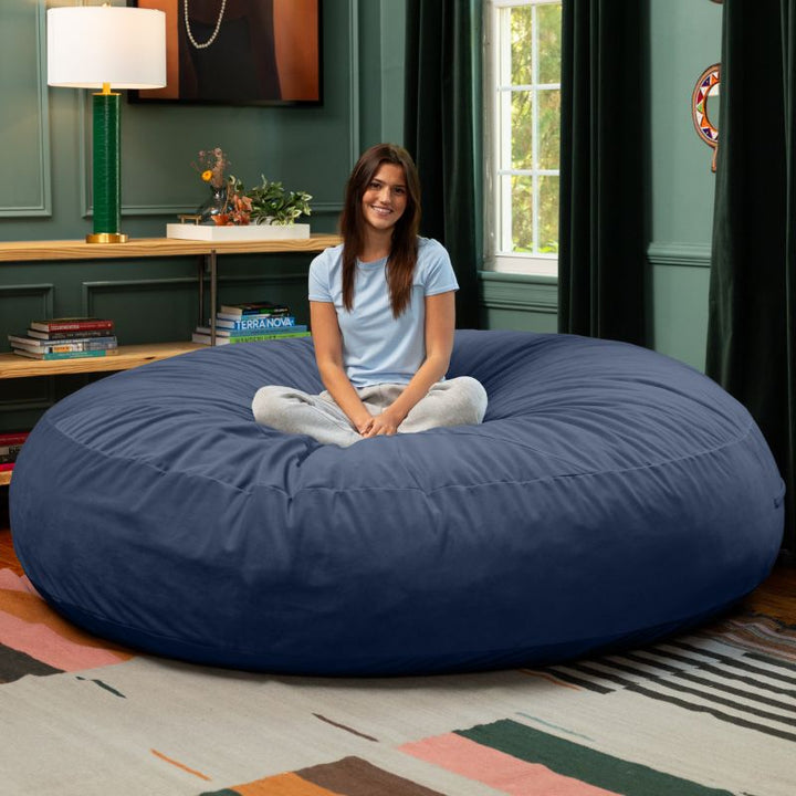 A woman sitting cross-legged on a blue Jaxx Cocoon 7ft XL bean bag chair, a calming oversized lounger