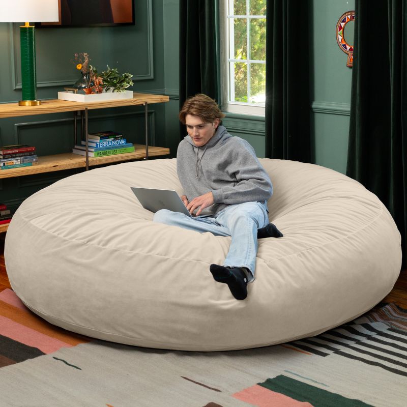 A man working on a laptop on a cream Jaxx Cocoon 7ft XL bean bag chair, a soft oversized sensory seat