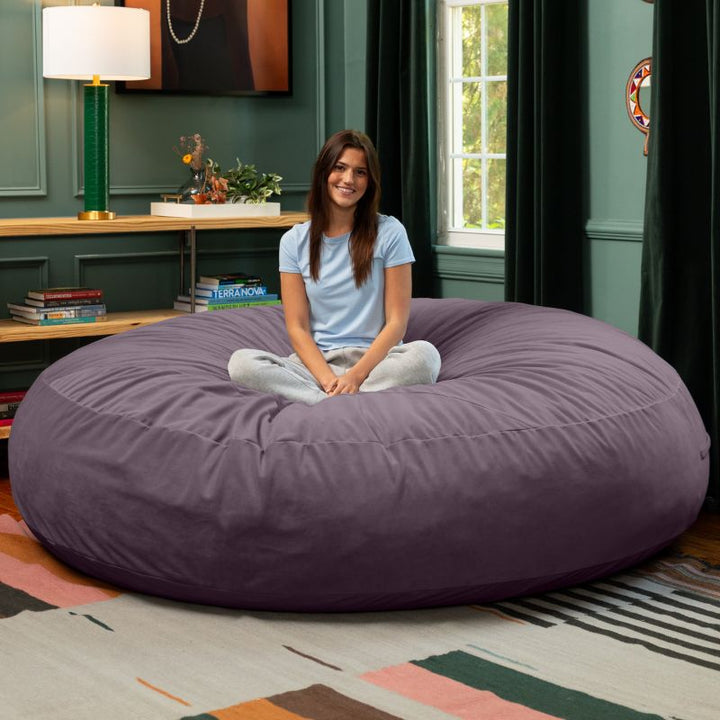 A woman sitting cross-legged on a purple Jaxx Cocoon 7ft XL bean bag chair, an extra-large sensory seat