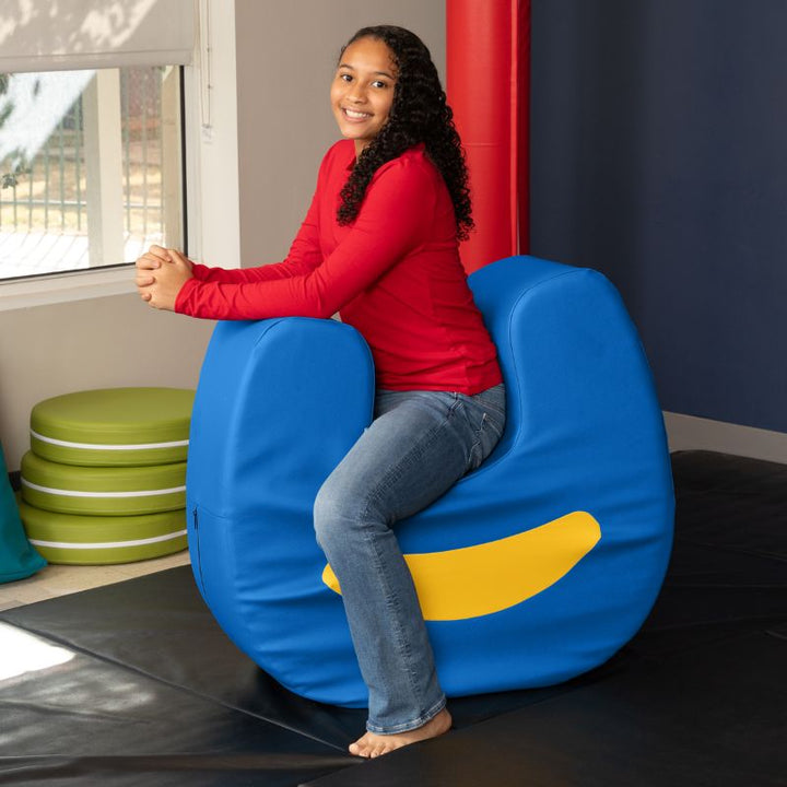 A child seated on the Jaxx Zipline Loveseat in a therapy room, with padded wall bolsters visible in the background