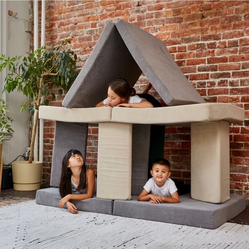 Three children playing in a large fort built from the Kiddie Couch in Grey and Beige, showing collaborative active sensory play