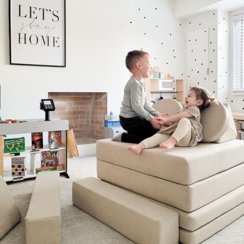 Two children stacking and playing on the Kiddie Couch cushions in Sand Beige, supporting proprioceptive input and active sensory play