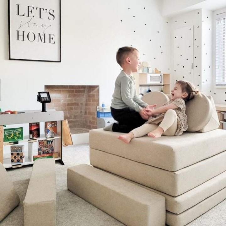 Two children stacking and playing on the Kiddie Couch cushions in Sand Beige, supporting proprioceptive input and active sensory play