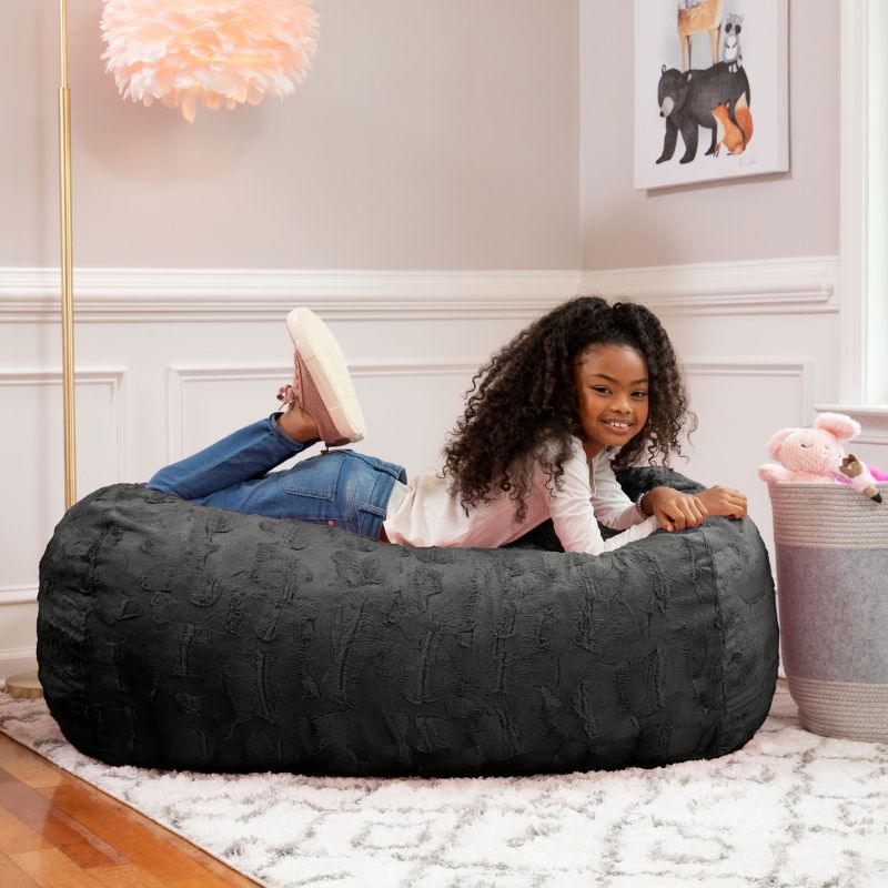 A girl relaxes on the Jaxx Lounger 4ft faux fur bean bag lounger for kids in Black, styled in a cozy bedroom setting.