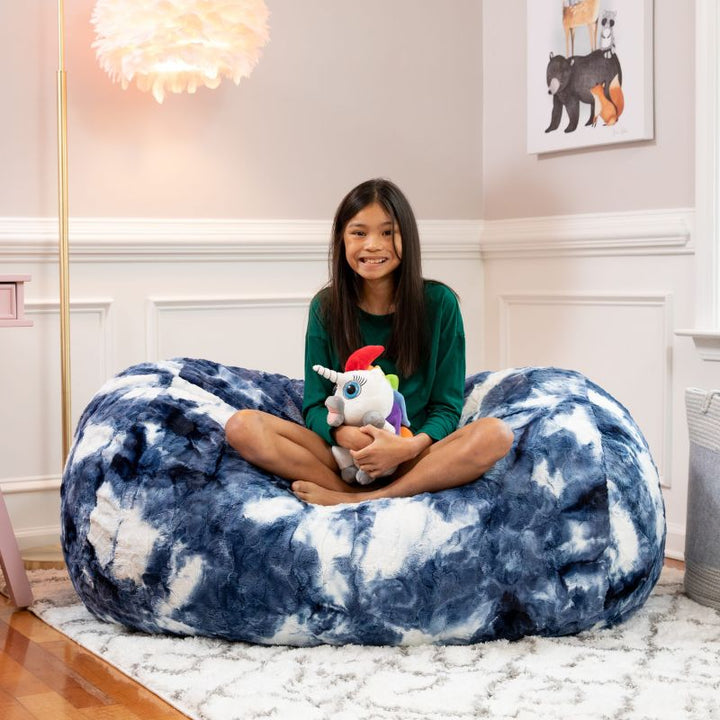 A girl sits with a toy on the Jaxx Lounger 4ft faux fur bean bag lounger for kids in Blueprint, a tie-dye sensory bean bag chair, styled in a bedroom setting.