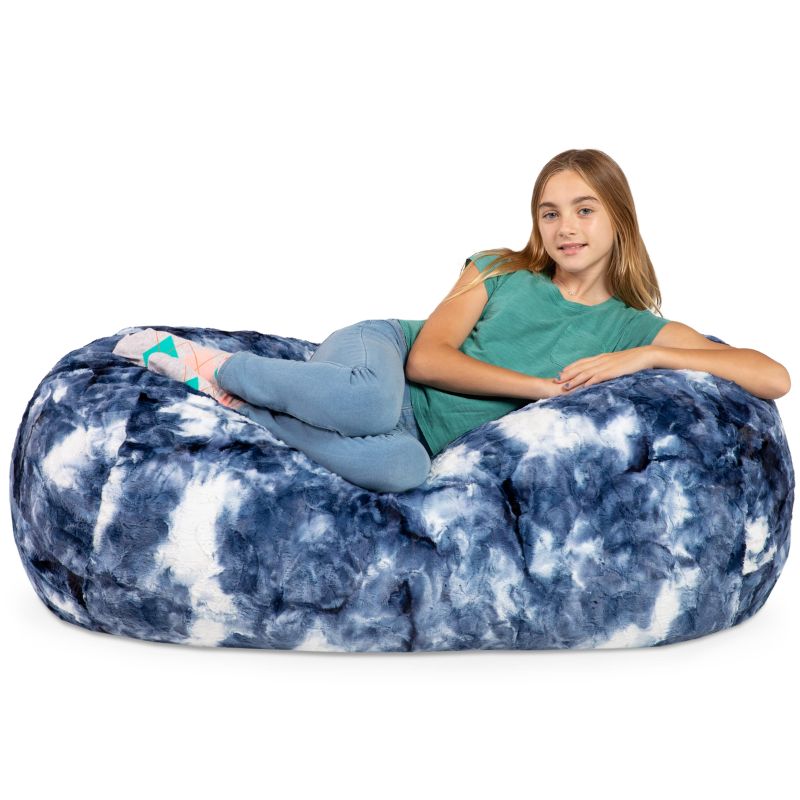 A girl reclines on the Jaxx Lounger 4ft faux fur bean bag lounger for kids in Blueprint, a tie-dye sensory bean bag chair.