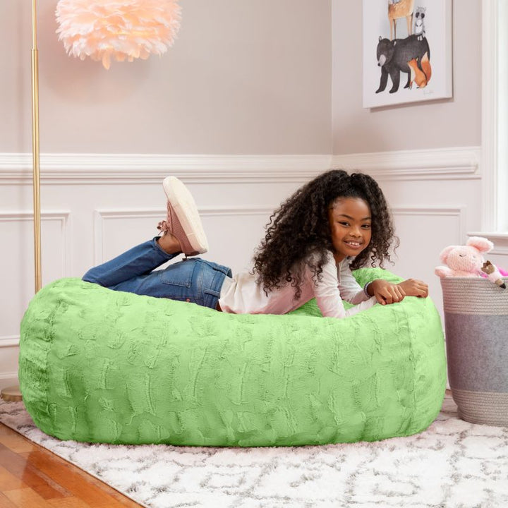 A girl relaxes on the Jaxx Lounger 4ft faux fur bean bag lounger for kids in Green, styled in a cozy bedroom setting.