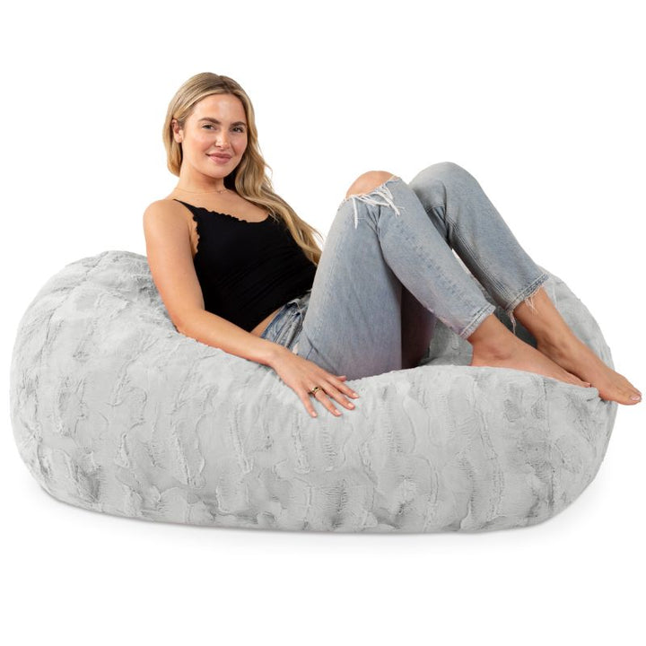 A woman sits on the Jaxx Lounger 4ft faux fur bean bag lounger in Grey, showing the kids bean bag chair sized for teens and adults too.