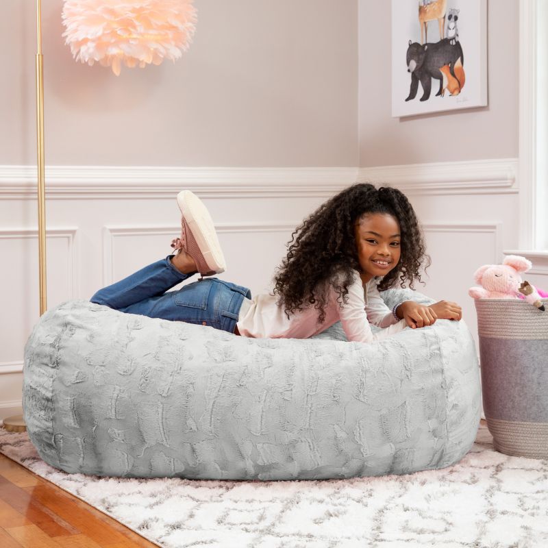 A girl relaxes on the Jaxx Lounger 4ft faux fur bean bag lounger for kids in Grey, styled in a cozy bedroom setting.