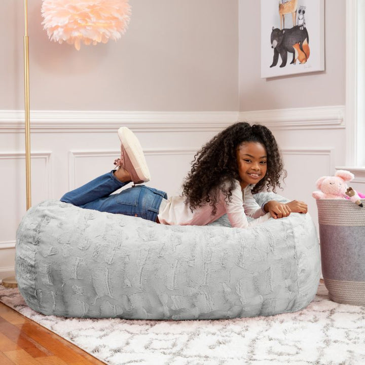 A girl relaxes on the Jaxx Lounger 4ft faux fur bean bag lounger for kids in Grey, styled in a cozy bedroom setting.