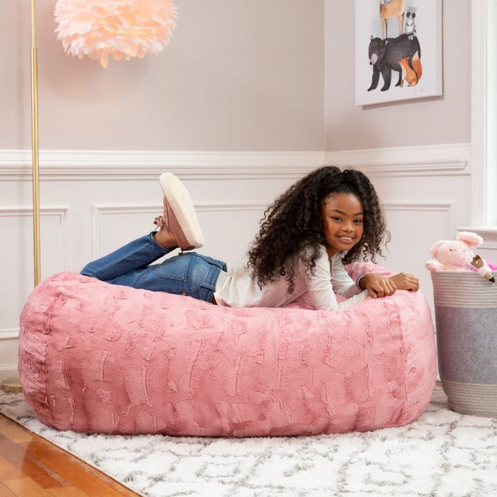 A girl relaxes on the Jaxx Lounger 4ft faux fur bean bag lounger for kids in Pink, styled in a cozy bedroom setting.