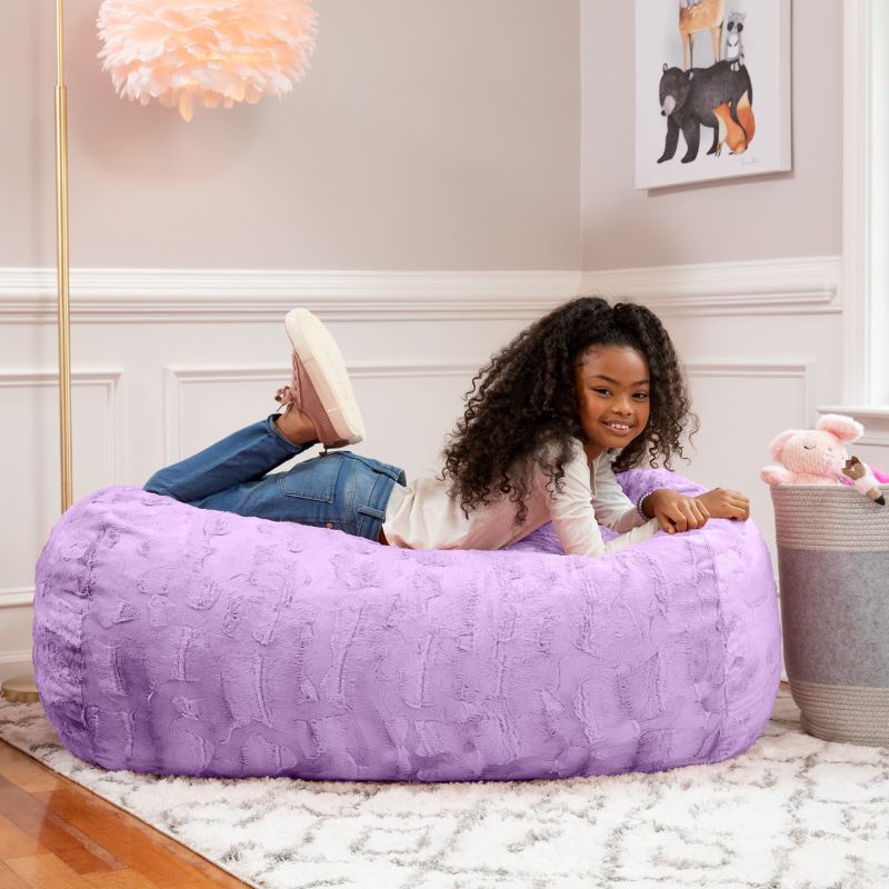 A girl relaxes on the Jaxx Lounger 4ft faux fur bean bag lounger for kids in Purple, styled in a cozy bedroom setting.
