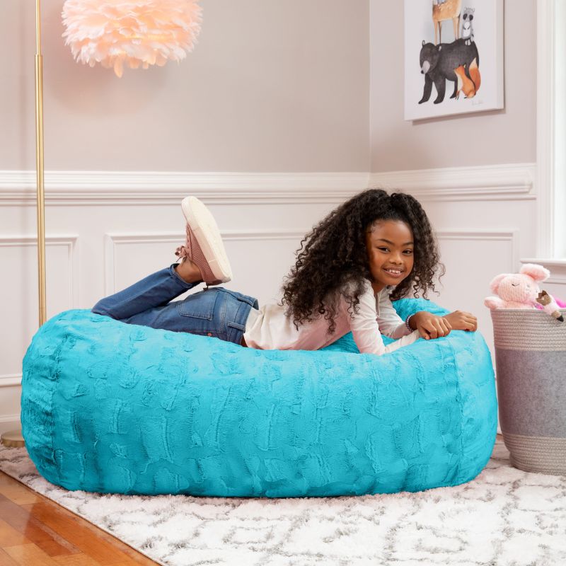 A girl relaxes on the Jaxx Lounger 4ft faux fur bean bag lounger for kids in Teal, styled in a cozy bedroom setting.
