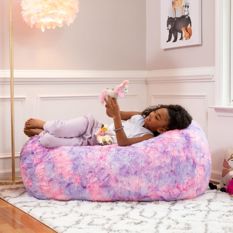 A girl plays with a toy while lying on the Jaxx Lounger 4ft faux fur bean bag lounger for kids in Unicorn, a pastel tie-dye sensory bean bag chair.