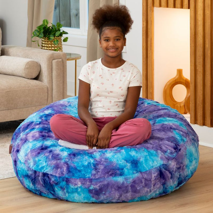 Child sitting cross-legged on the Raven-Symoné Lil' Cuddler kids bean bag lounger in Lush Tie-Dye Fur Mystic Blue & Purple