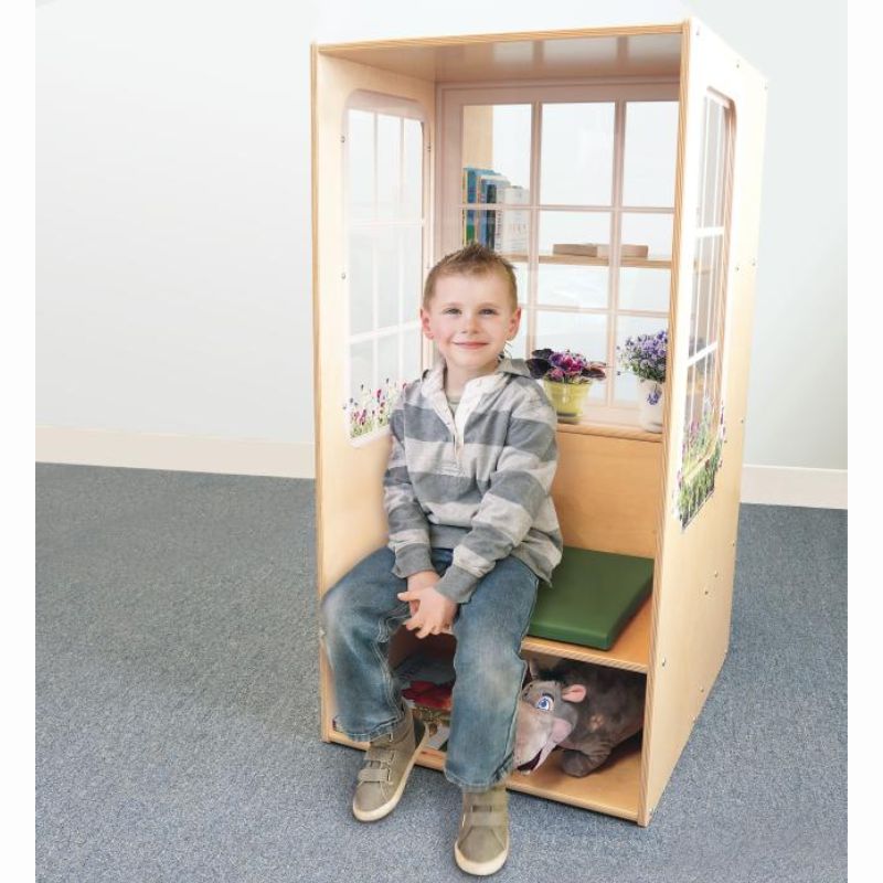 Smiling child sitting on the green cushion of the Whitney Brothers Nature View Window Seat, a kids reading nook with books on the rear shelves and a plush toy tucked in the under-seat storage