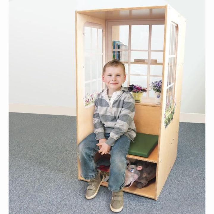 Smiling child sitting on the green cushion of the Whitney Brothers Nature View Window Seat, a kids reading nook with books on the rear shelves and a plush toy tucked in the under-seat storage
