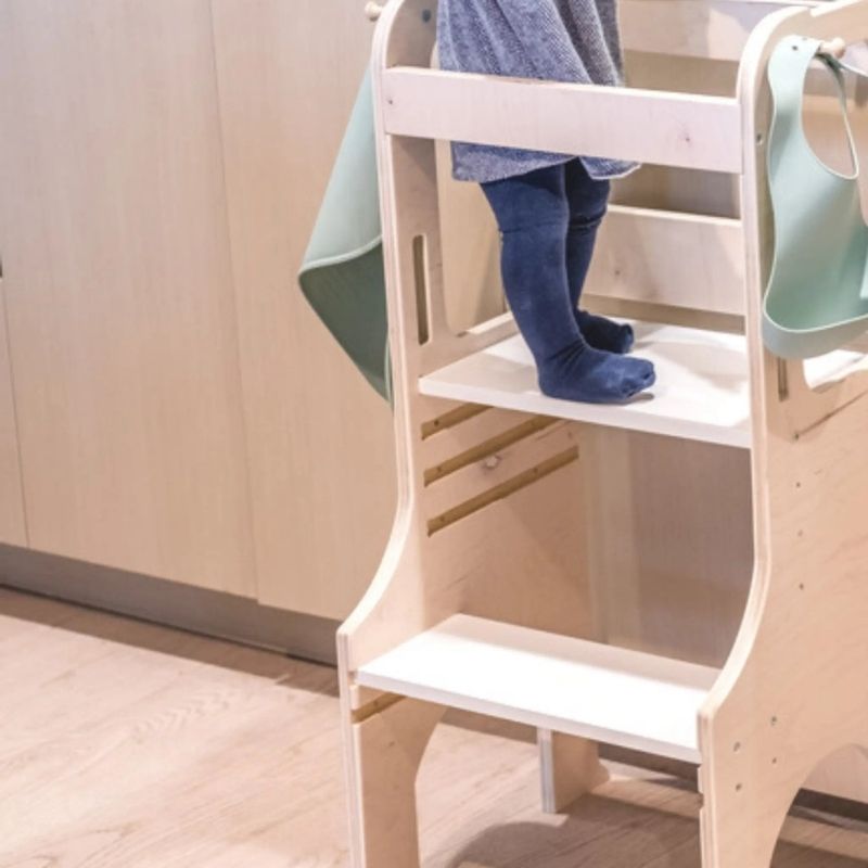 Close-up of child standing safely in the Sapiens Child Kiwi Kitchen Tower, showing the enclosed rail design and adjustable platform height supporting toddler independence at counter level