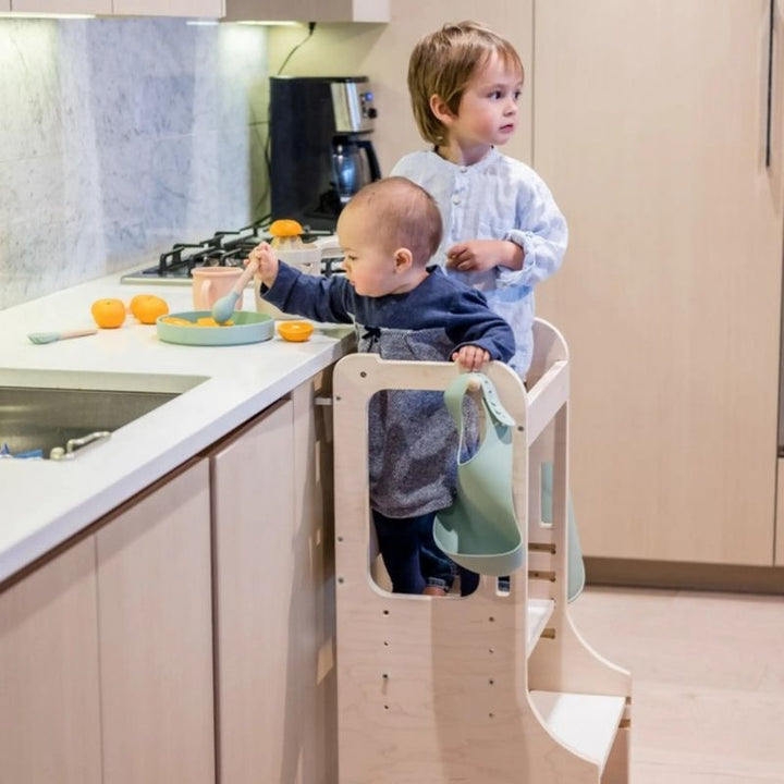 Two children standing in Sapiens Child Kiwi Kitchen Towers side by side, showing the learning stool supporting sibling participation in cooking and kitchen activities