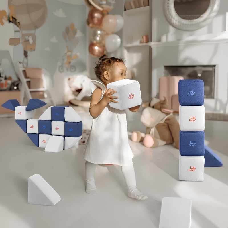 A toddler playing with the JollyHeap Mini magnetic block set in a nursery, building a whale shape from soft fabric-covered cubes and triangles
