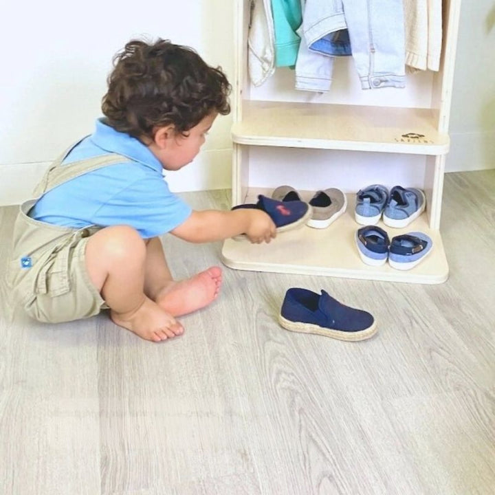 Child actively putting on shoes at the Sapiens Child Mango Montessori Cubby Coat Rack, showing the child-sized seat that supports independent shoe management as part of the daily dressing routine
