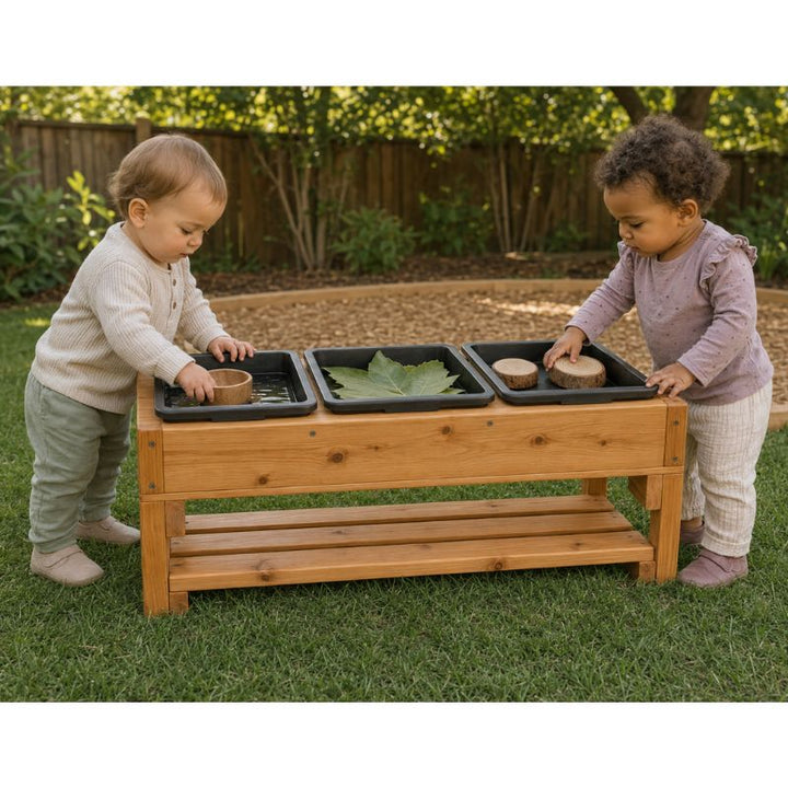 Two toddlers standing at the ONP 3 Bin Sensory Table outdoors, touching water, a large leaf and wood slices across the three bins