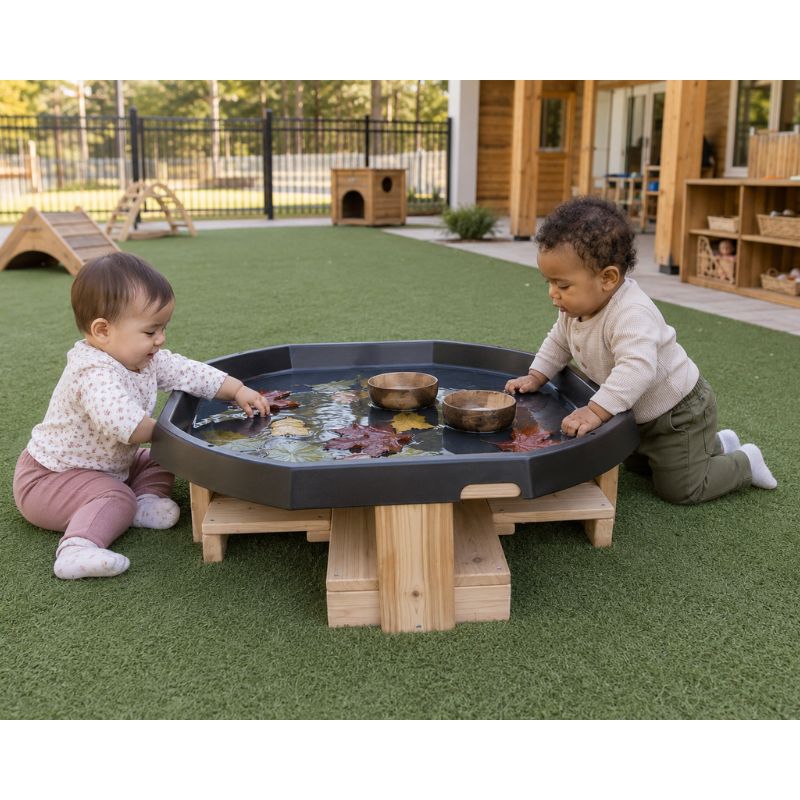 Two toddlers kneeling at the ONP Active World Tray Stand on turf, touching floating leaves and wooden bowls in the water tray