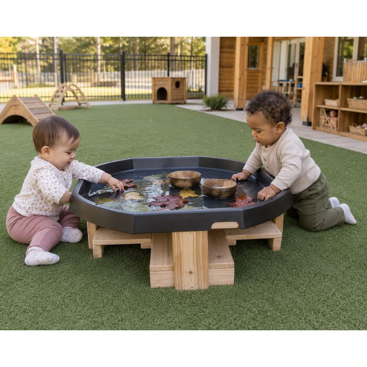 Two toddlers kneeling at the ONP Active World Tray Stand on turf, touching floating leaves and wooden bowls in the water tray