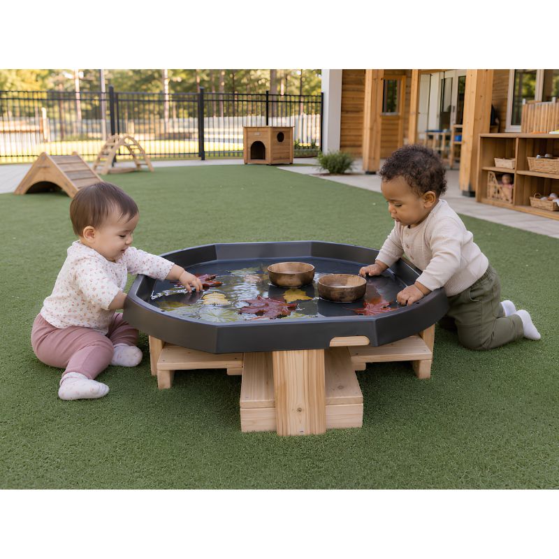 Two infants seated at the low ONP Infant Active World Tray Stand outdoors, floating leaves and wooden bowls in the water-filled tray
