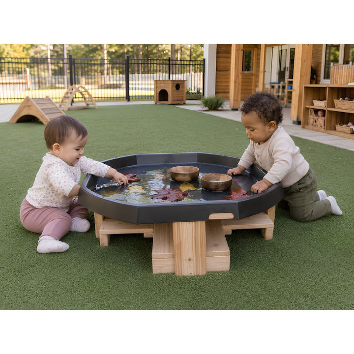 Two infants seated at the low ONP Infant Active World Tray Stand outdoors, floating leaves and wooden bowls in the water-filled tray