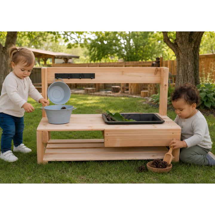 Two toddlers playing at the low ONP Infant Mud Kitchen outdoors, one lifting a hanging bucket and one scooping pinecones from a bowl