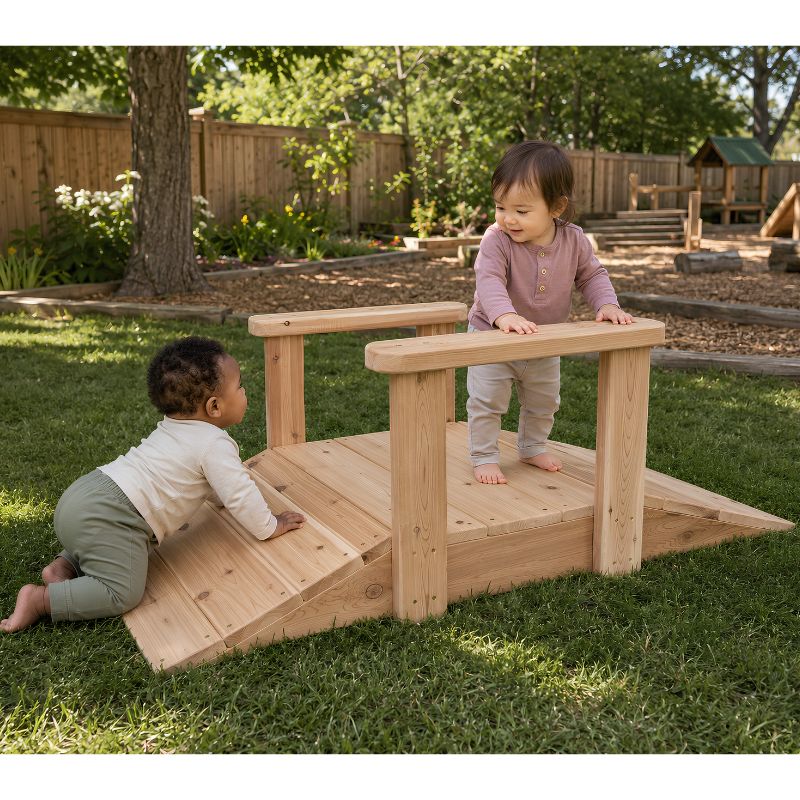 An infant crawling up the wooden ONP Infant Ramp outdoors while a toddler stands at the top holding the handrail